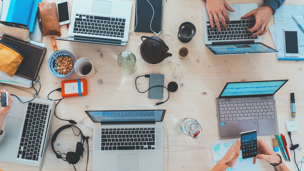 technology like computers, phones, charges, headphones and more on a table shared between coworkers