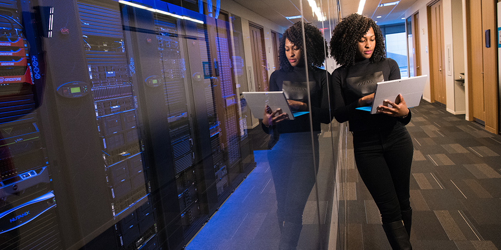 Woman leaning on a glass wall separating her from wall of data servers managed by AI.