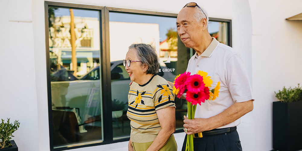 Senior real estate with two senior citizens walking around outsides holding flowers.