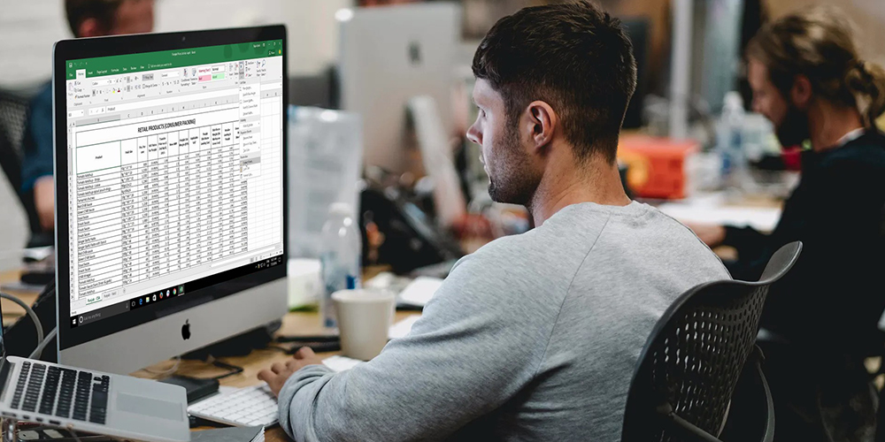 Spreadsheets open on desktop with man in office setting.