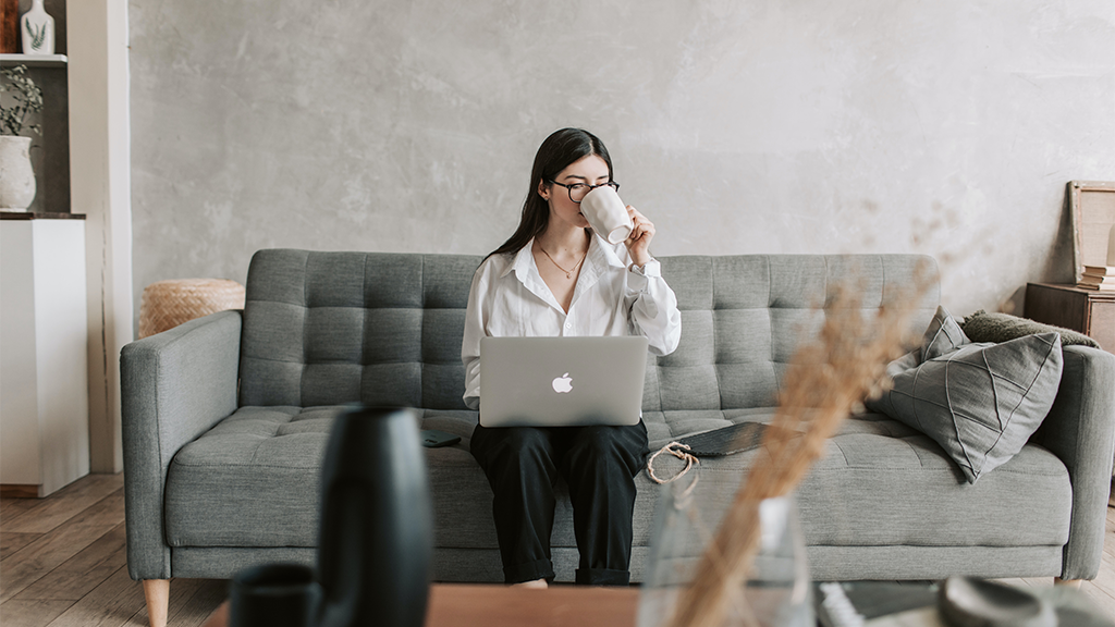 A woman seated on a modern couch sipping coffee and typing out emails on her laptop.