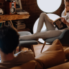 A man sits in the foreground working on his laptop while a woman sits off to the side reading a book. Both people are inside a comfortable, warmly lit bohemian apartment.