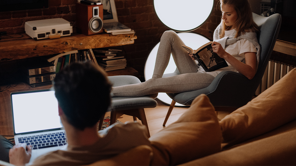 A man sits in the foreground working on his laptop while a woman sits off to the side reading a book. Both people are inside a comfortable, warmly lit bohemian apartment.