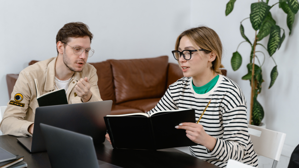 A man and a woman sitting together and talking at a table, while the woman holds up her notebook to show her notes, talking about disclosure.