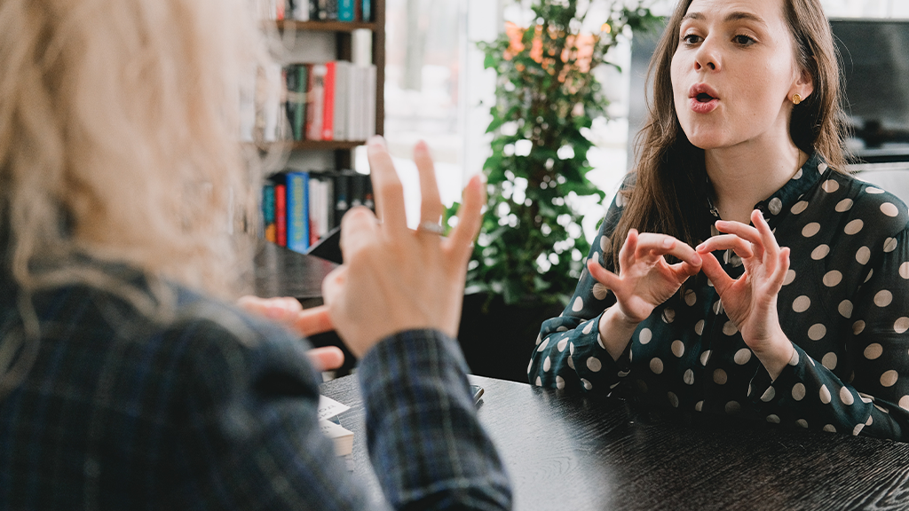 A woman practices ASL (American sign language) with another person, speaking to emphasize what they're saying.
