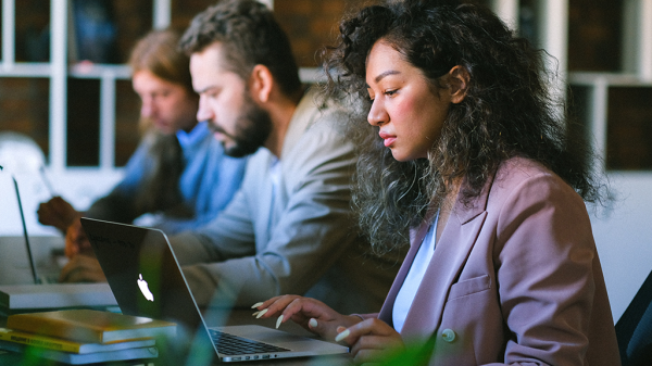 A line of people seated at a desk with their laptops open in front of them, working seriously in light of overemployment.