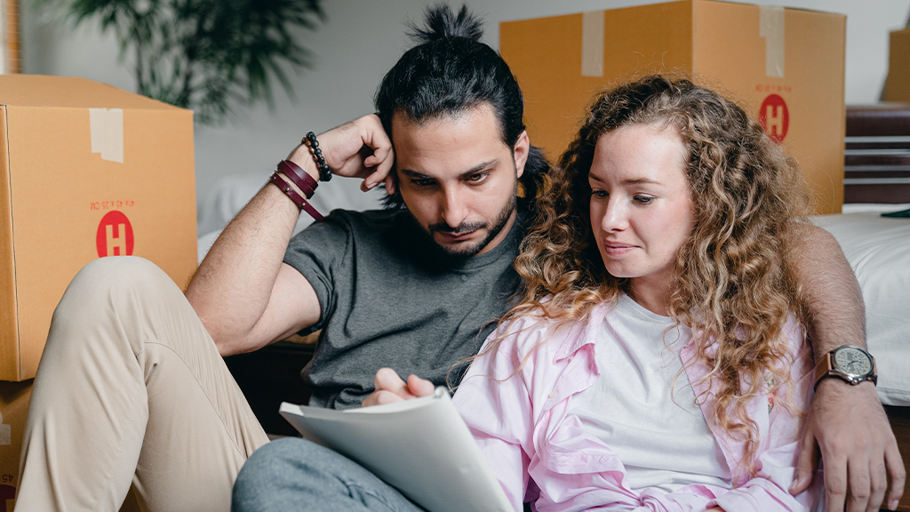A man and a woman surrounded by moving boxes reviewing documents with contemplative expressions.