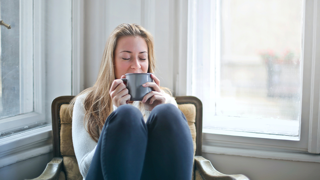 A white woman relaxes in a seat in a corner by some windows, managing stress by sipping on a cup of a nice beverage.