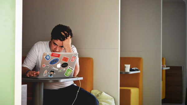 A man sits in a booth in a cafe, working on a laptop with a stressed expression as he fights comparison