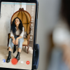 A phone showing a woman being recorded for a Pinterest short form video. She wears bohemian clothes and big black boots and sits in a wicker chair.