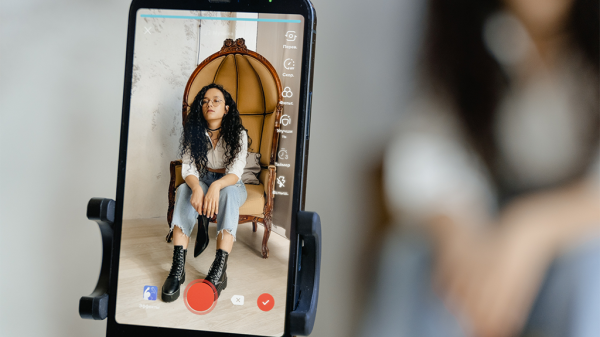 A phone showing a woman being recorded for a Pinterest short form video. She wears bohemian clothes and big black boots and sits in a wicker chair.