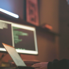 A woman with blue hair sits at a laptop with two larger desktop monitors on the desk with various code information on it to prepare for cybercrimes.