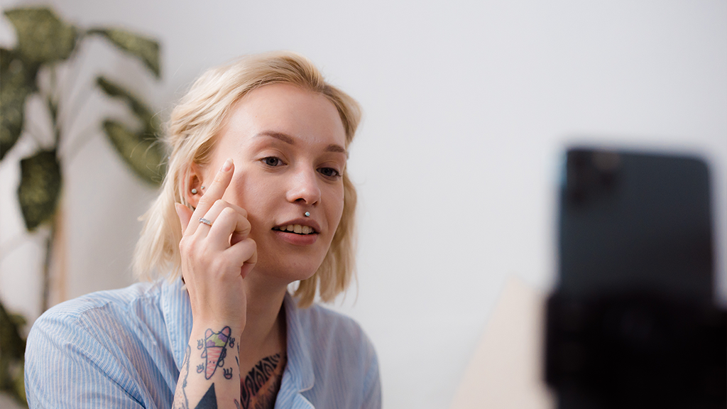 A woman with tattoos on her arm points to her face as she records in front of her phone for a Tiktok video.