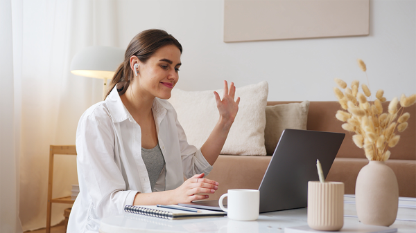A woman waves at the laptop with airpods in as she checks in for daily stand-ups.