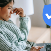 A woman sits on a couch with a laptop open, browsing with a thoughtful expression. A blue verified check mark in a blue shield shape sits on top of the scene.