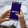 A woman holds a phone open to Mastodon app while she sits on a white bed.