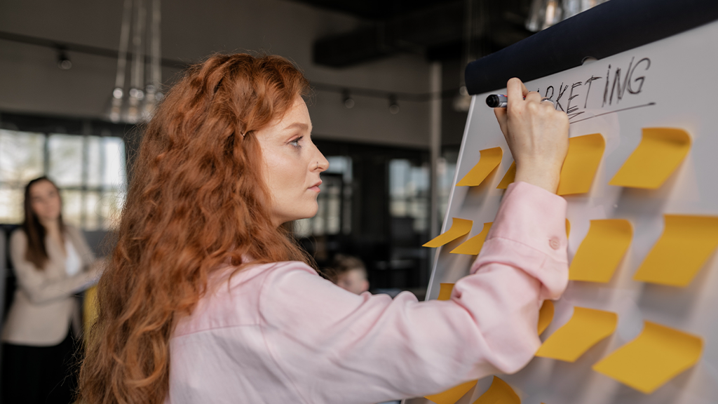 A woman with long red hair stands at a marketing board with multiple post-it notes, manually writing what AI services can assist with.