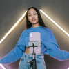 A woman stands in front of a phone on a phone stand gesturing welcomingly, with purple and yellow neon bars on the wall behind her while she records for Tiktok.
