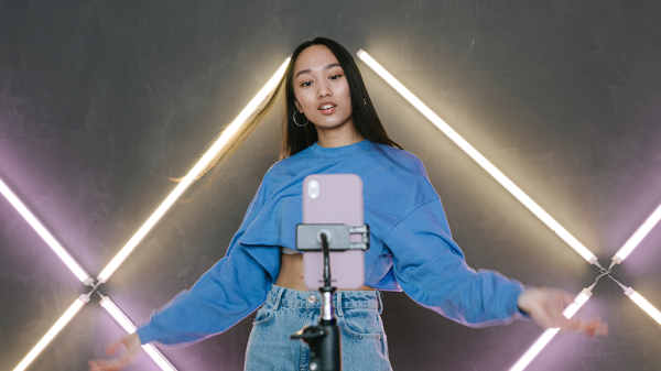 A woman stands in front of a phone on a phone stand gesturing welcomingly, with purple and yellow neon bars on the wall behind her while she records for Tiktok.