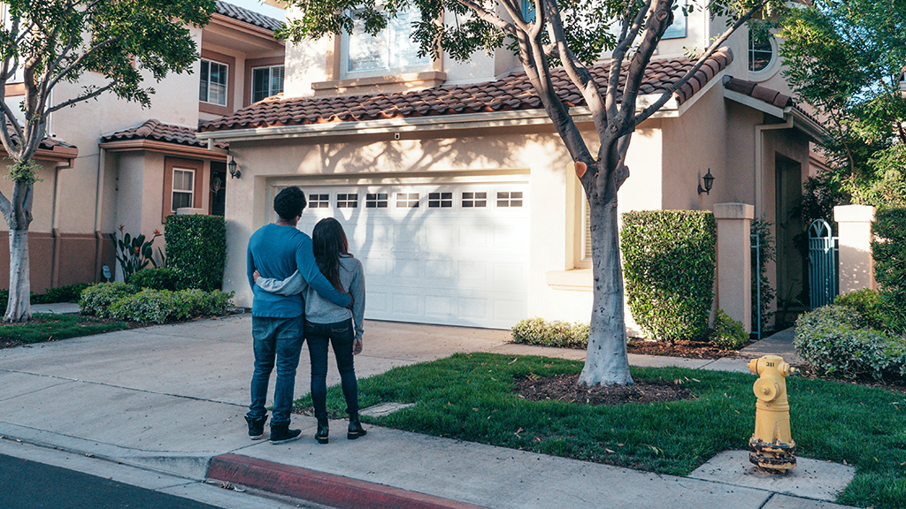 A young couple holding each other as they stand outside of a house pondering homeownership.
