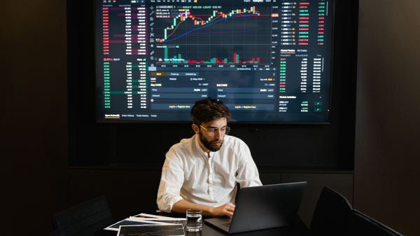 A man sits in a meeting room with a laptop and a large digital screen of charts and data on the housing market behind him.