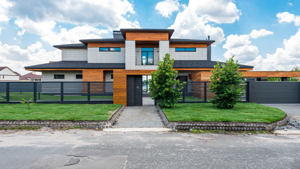 A large affluent suburb home with multiple stories and a black metal fence surrounding the property. A well manicured lawn and shrubs sit outside.