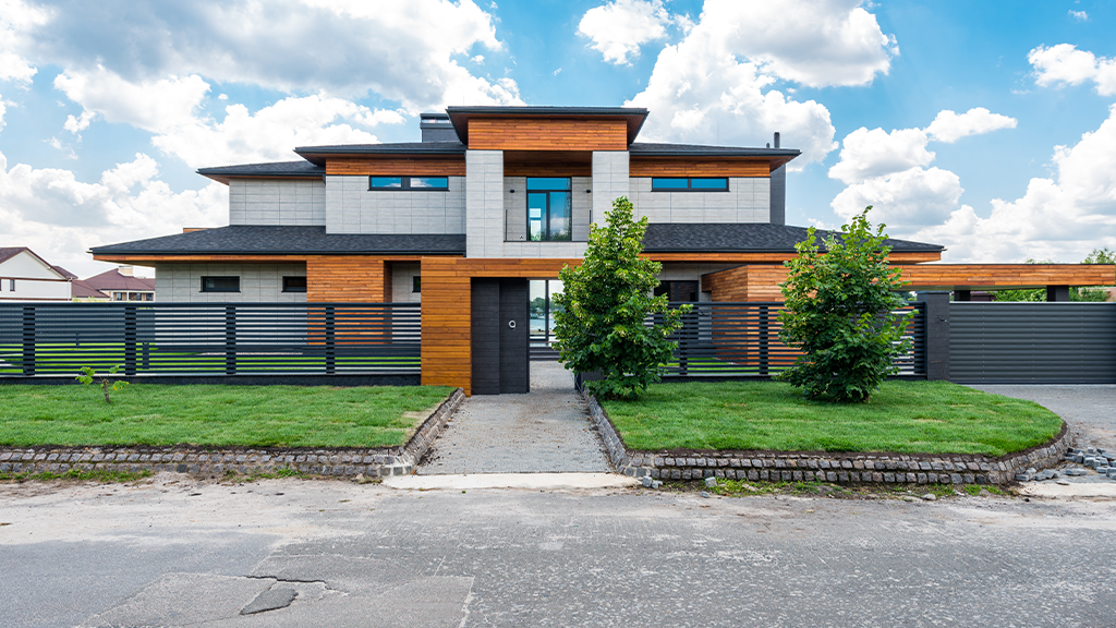 A large affluent suburb home with multiple stories and a black metal fence surrounding the property. A well manicured lawn and shrubs sit outside.
