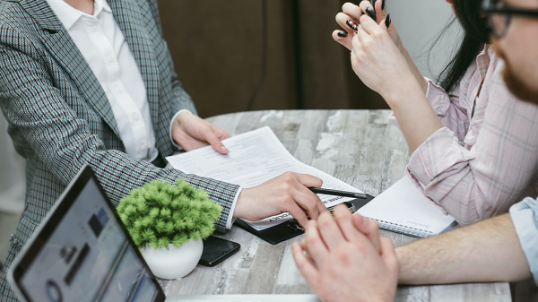 A side view of three people gathered around a table, from the neck down. Mortgage paperwork is on the table and a person in a suit is reading it to the others at the table.