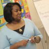 A Black female teacher stands at the front of a classroom with teaching tools around her smiling at her class.