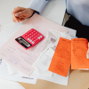 A table of reciepts, white and orange papers relating to homeownership, and a person using a pink calculator in a top down view of their table.