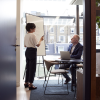 A view through an office door of a woman standing and giving a presentation to a man sitting in a desk.