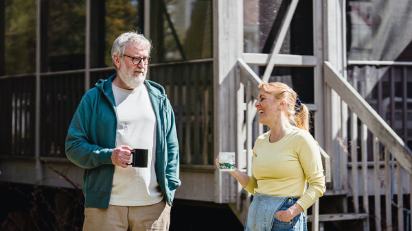 A pair of seniors, a man and a woman, standing outside of a home with mugs of a drink in their hand, laughing as they talk.