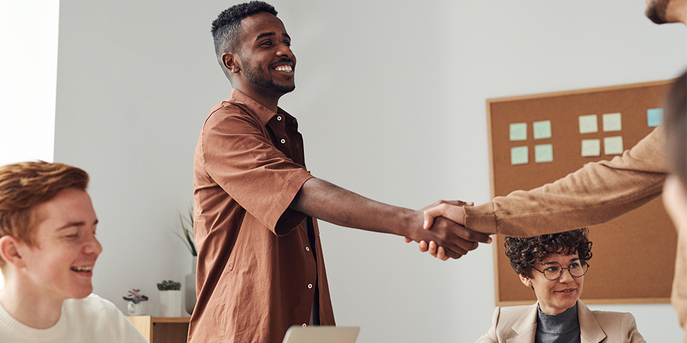 Two men standing in meeting room with others, shaking hands as they agree to be co-founder together.