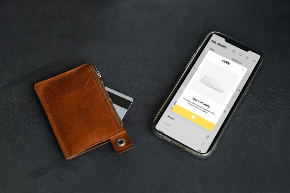 Brown leather wallet with tip of credit card sticking out next to a iPhone showing a shoe purchase on the Nate App. 