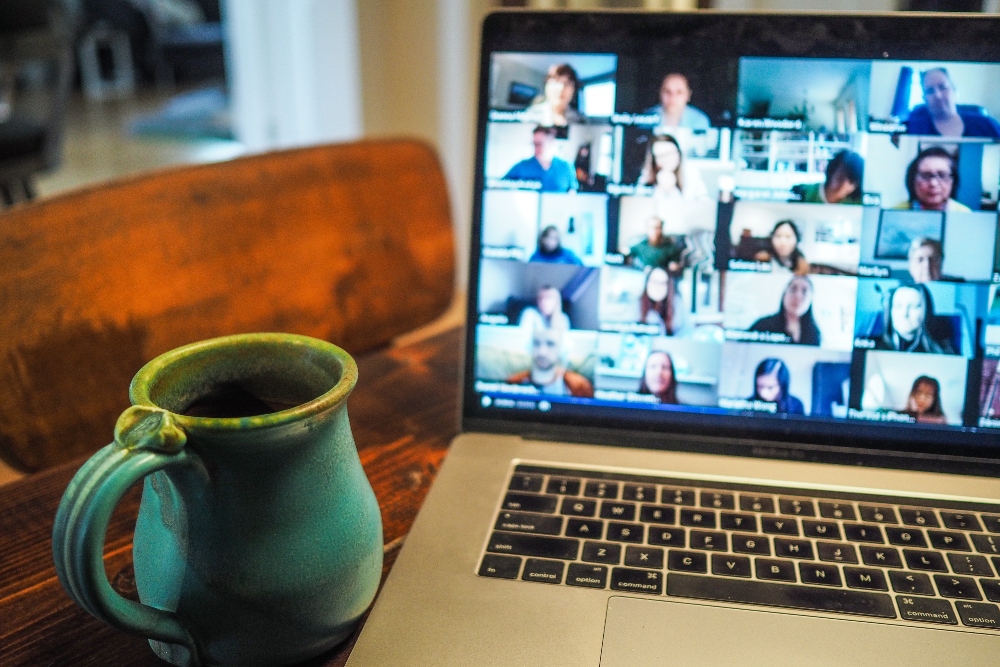 Multi-member Zoom call on a Apple Mac laptop with a blue mug of black coffee next to it.