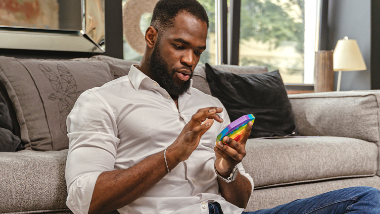 A Black man sits on the floor in front of a couch with a rainbow pop toy in his hand. He is smiling and enjoying his adult toys.