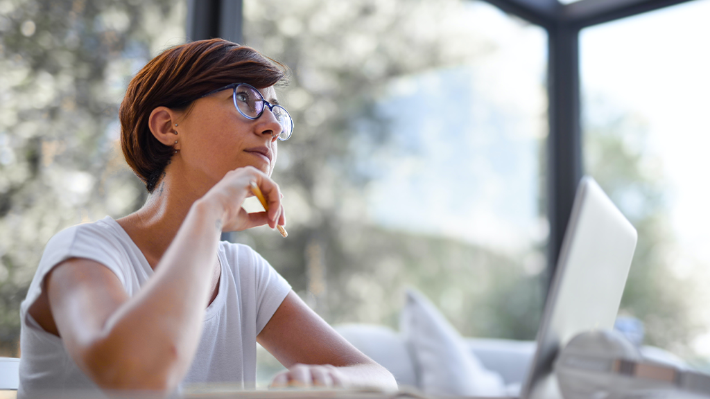 A woman sits in a well lit room, thinking about business as she sits at a laptop with a contemplative expression.
