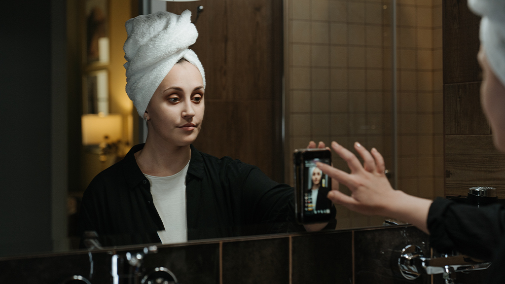 InstagramVideo A woman with her hair tied up in a towel to dry taps on her phone stuck to a bathroom mirror, recording an Instagram Reels video