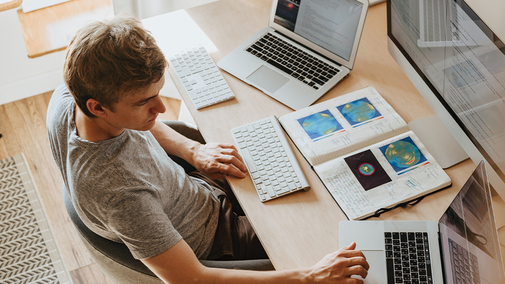 A man sits at a desk with two open laptops, a desktop, and a book open as he checks his ChatGPT notes.