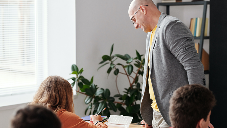 A bald man stands over a student in a classroom, helping with teachers resources and knowledge.