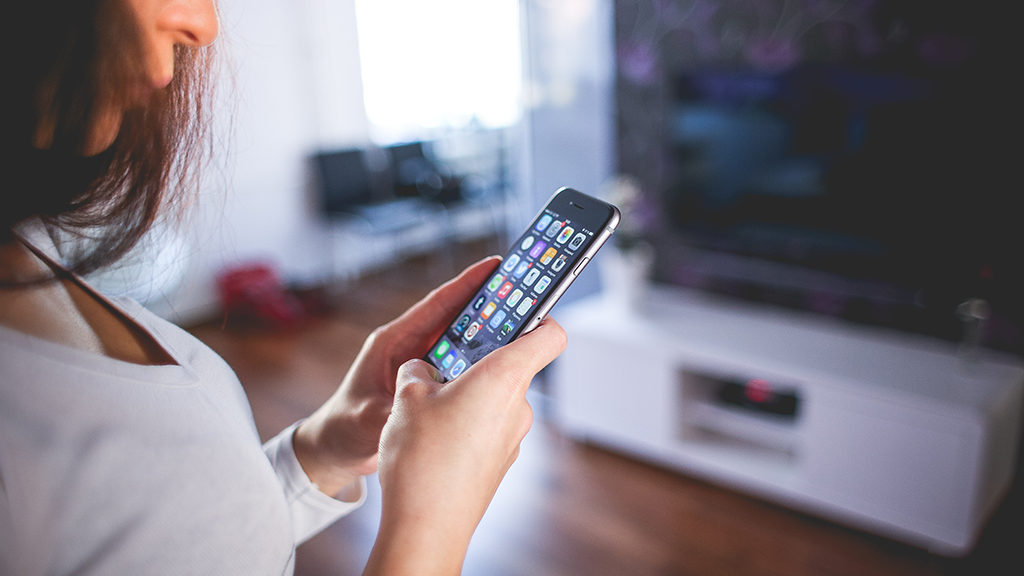 A person partially cut off and holding up an Apple phone with various sherlocked apps open. They stand in a living room with a turned-off TV in the blurred background.