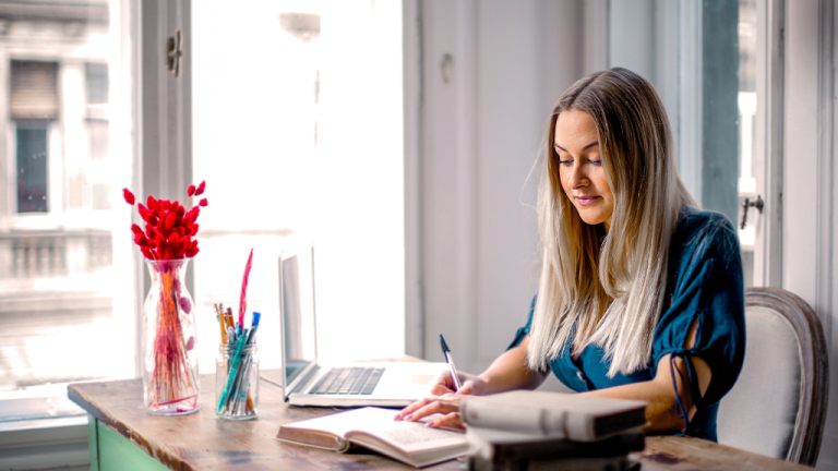 A woman seated at a desk inside of an office writing in a notebook with a laptop open beside her in the return to office.