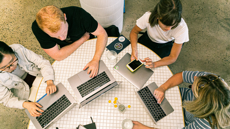A top-down view of a group of startup workers working at a round table and typing on laptops.