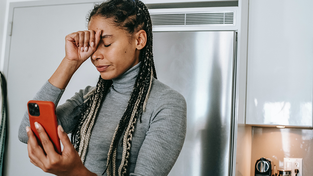 A woman holds a hand up in stressed position on her forehead while she looks at her phone with spam calls.