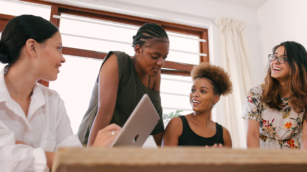 A group of women working together and standing over a table as part of the labor force, chatting animatedly.