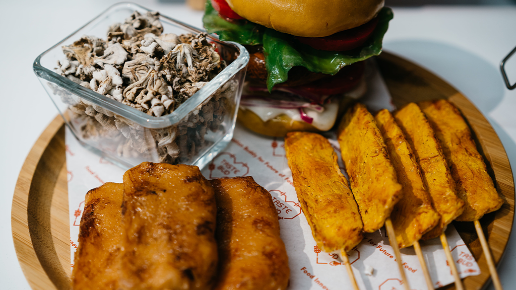 A plate of plant-based food including a skewer, a burger, and a bowl of mushrooms.