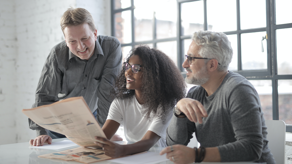A small group of employees, two older men and one younger Black woman, gathered around paperwork about AI and discussing earnestly.