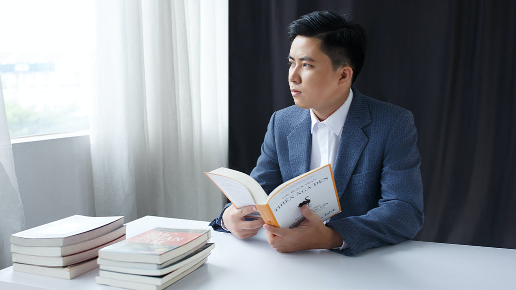 A business man with a book open on his desk looking thoughtfully out a window as he reads about efficiency.
