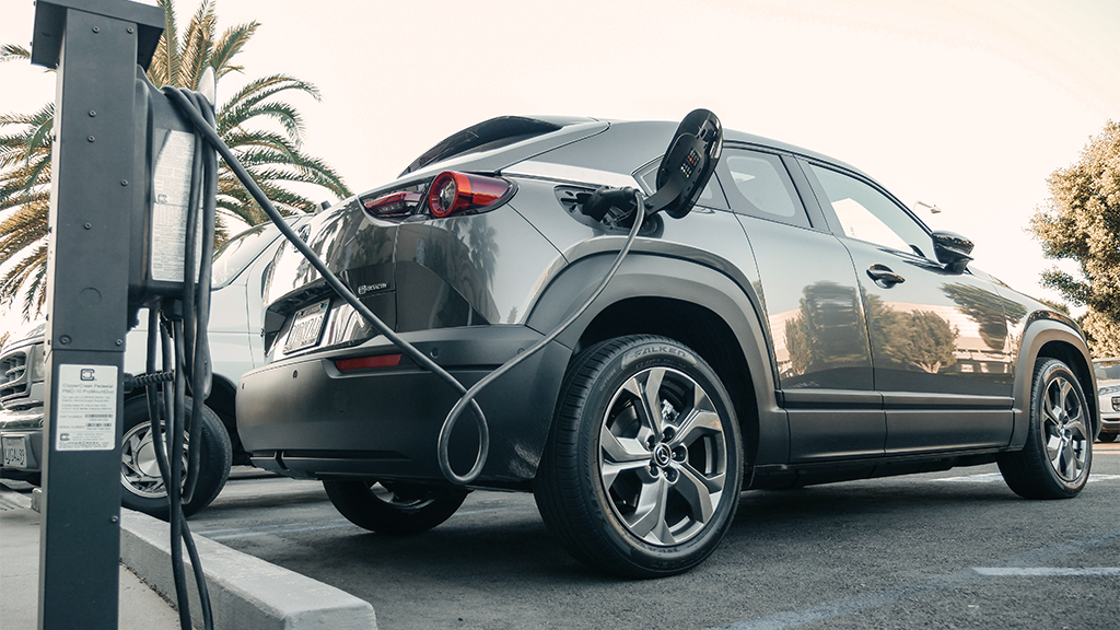 An electric vehicle hooked up to a charging station in a parking lot, with trees in the background.