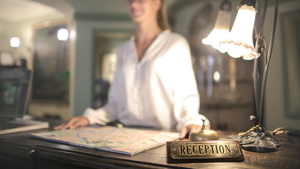 A woman standing at a hotel desk, with the focus on the foreground with the sign 'reception' illuminated by a frilled white desk lamp. The desk shows no ADA accommodation for the receptionist.